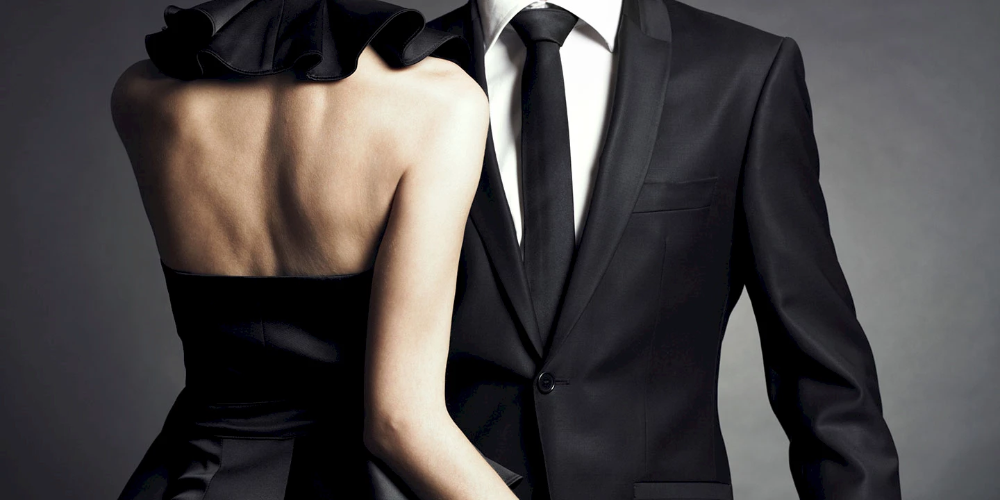 Eine Frau in einem schwarzen schulterfreien Abendkleid von hinten, ein Mann in einem schwarzen Anzug mit Krawatte von vorne. Von beiden sieht man nur die Oberkörper.