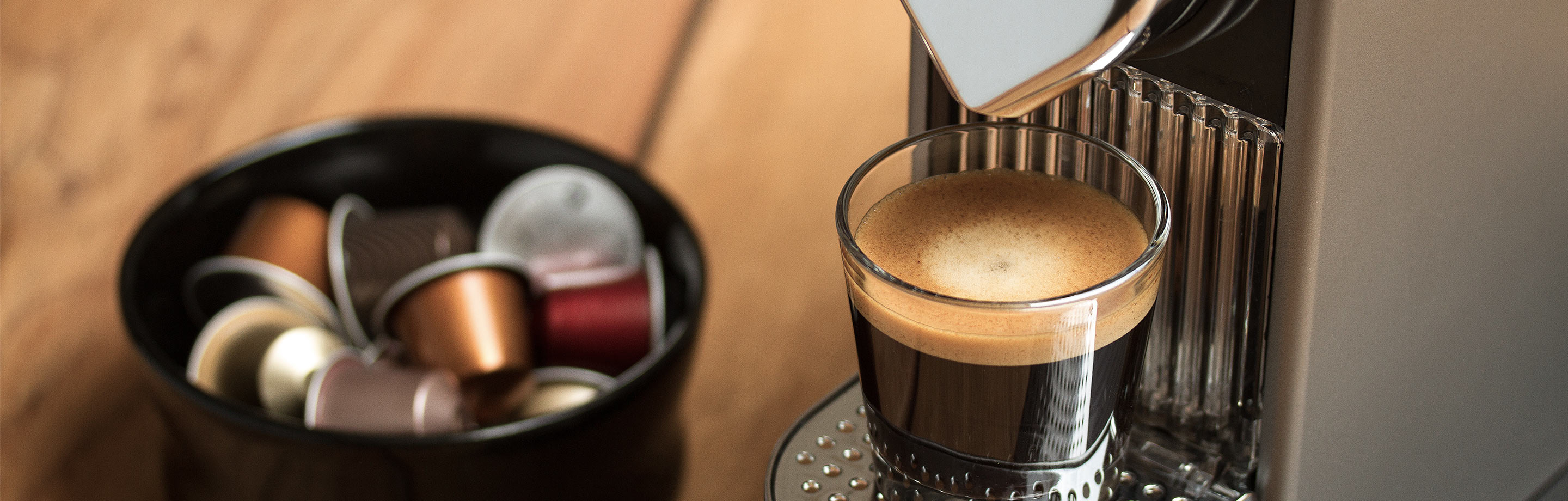 Eine Nespresso-Maschine mit einem gefüllten Espresso-Glas. Im Hintergrund befinden sich verschiedene Kaffeekapseln.
