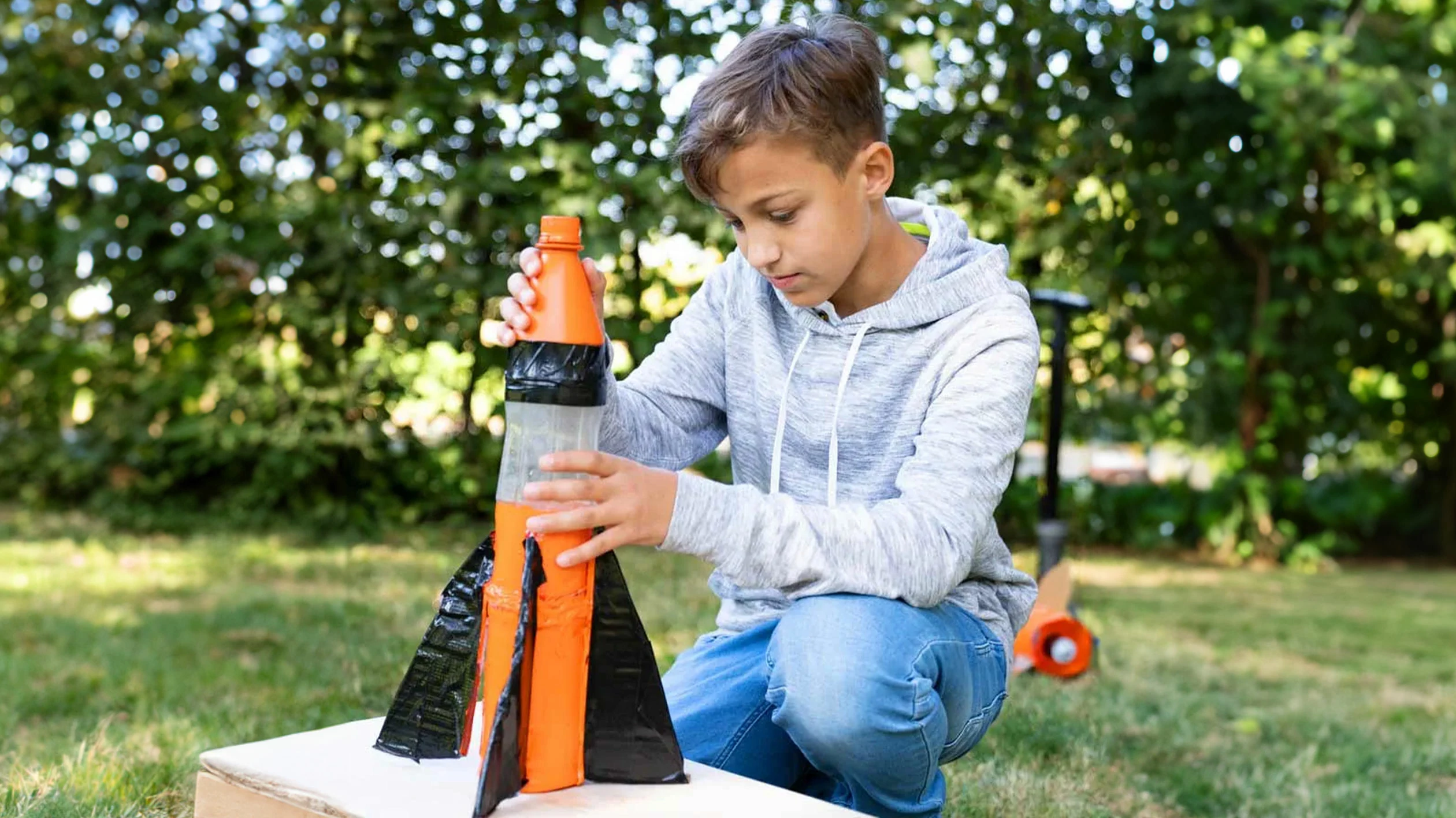 A boy kneels in the grass and tinkers with a children's toy in the shape of a rocket.