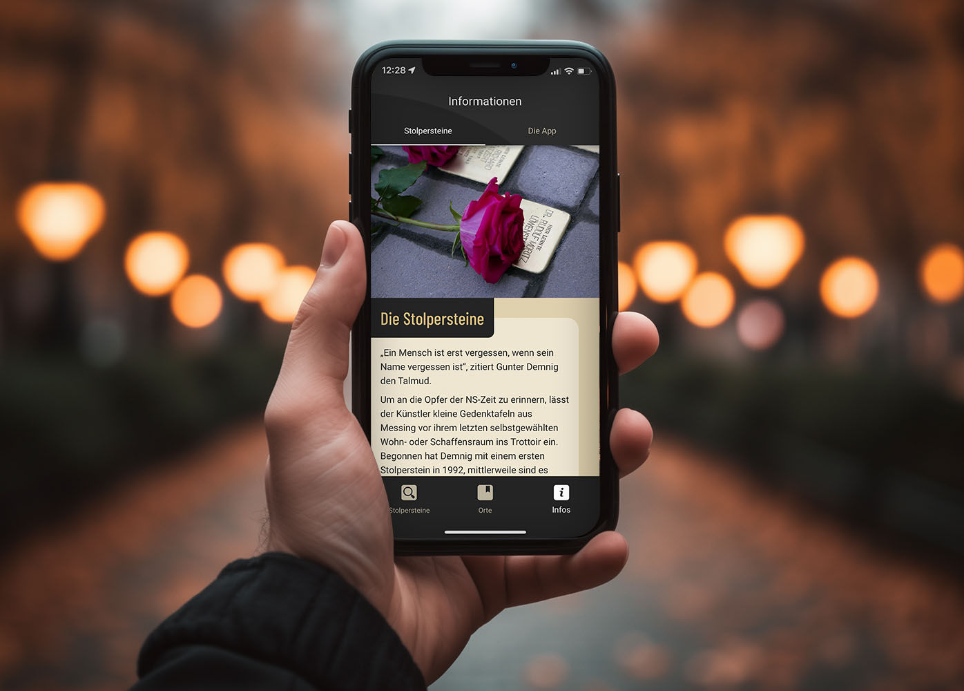 A hand holds a smartphone in front of a street background. On it is a picture of a stumbling stone with a red rose on it and an explanatory text about the app.