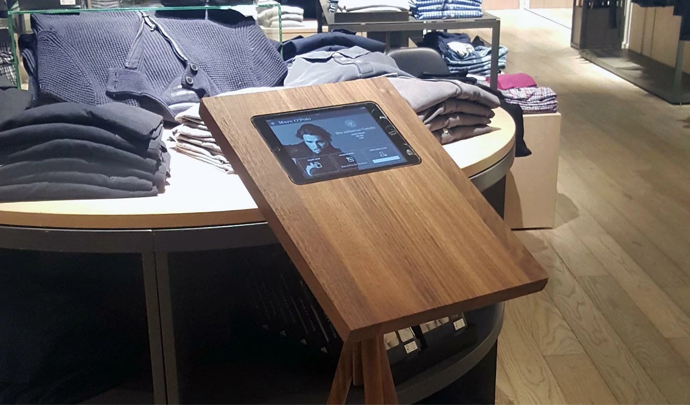 Scenery in a Marc'O'Polo store: Various items of clothing lie on a table. There is a wooden holder with an embedded tablet showing the sales app