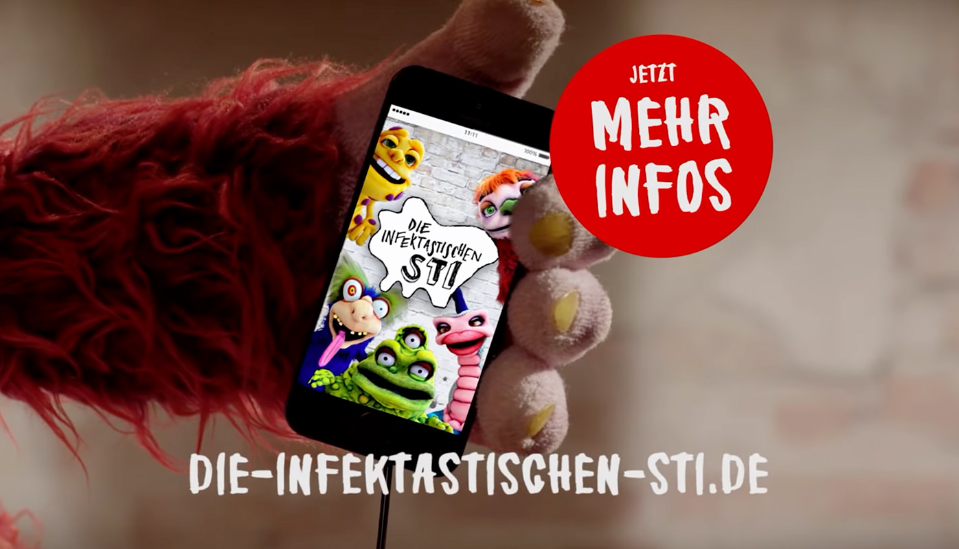 Ein Handy, auf dem die fünf Puppen der infektastischen STI abgebildet sind. Das Handy wird von der Hand einer Puppe gehalten. Auf dem Bild wird auf die Website die-infektastischen-sti.de dargestellt.