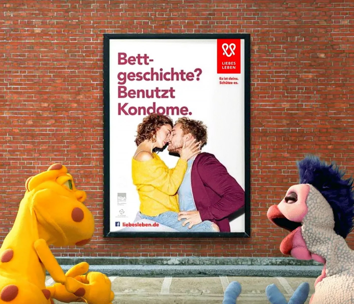 In the foreground, two puppets of the Infectious STI are talking. In the background is a wall with a poster from the Love Life campaign with the inscription "Bettgeschichte? Use condoms". On the poster, a man and a woman are kissing.