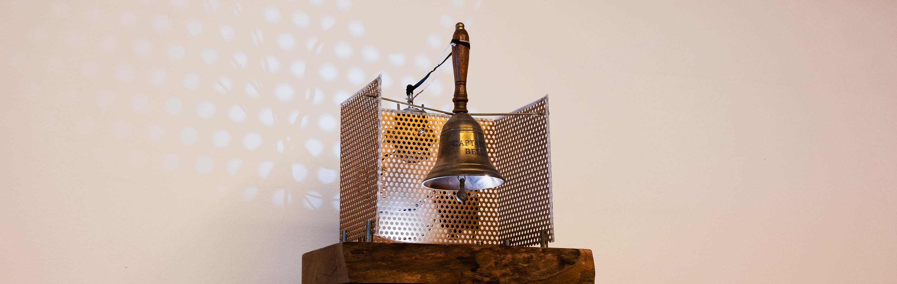 Eine alte Seemannsglocke aus Messing mit einem Holzgriff auf einem hölzernen Podest, auf der die Worte „Beer Captain“ eingraviert sind. Die Glocke ist mit einem Motor verkabelt.