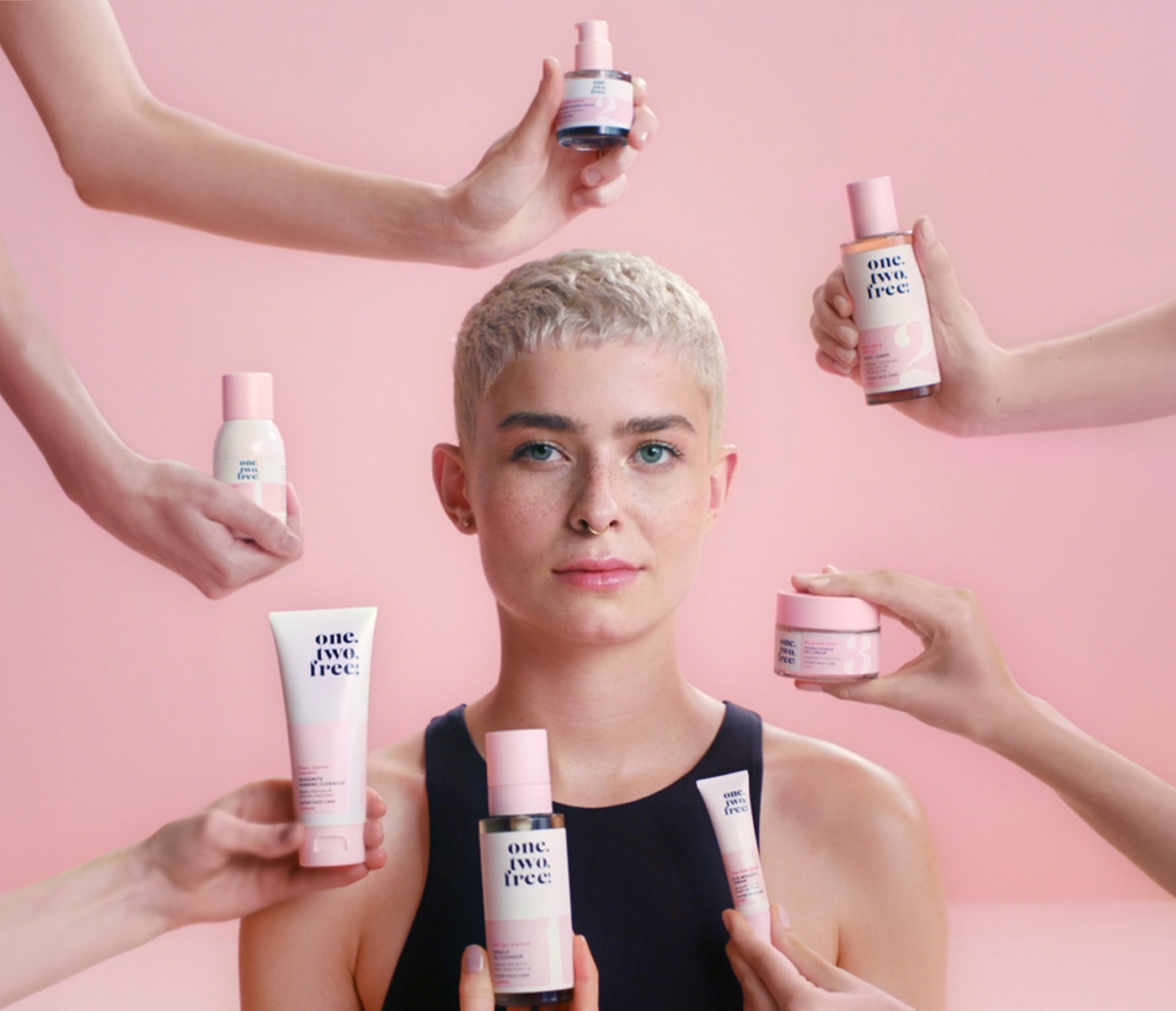 A female model in front of a monochrome background. Arms and hands with products protrude into the picture from all directions.