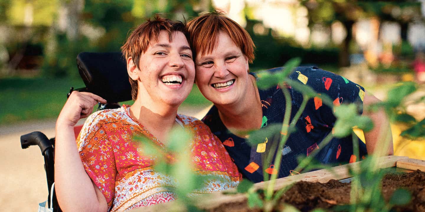 Portrait of two women in a green outdoor area. They lean their heads close together and laugh towards the camera.