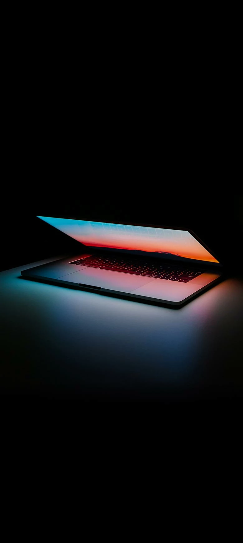 Ein Laptop vor dunklem Hintergrund. Das Display ist halb heruntergeklappt, dessen bunter Lichtschein die Szenerie beleuchtet. 