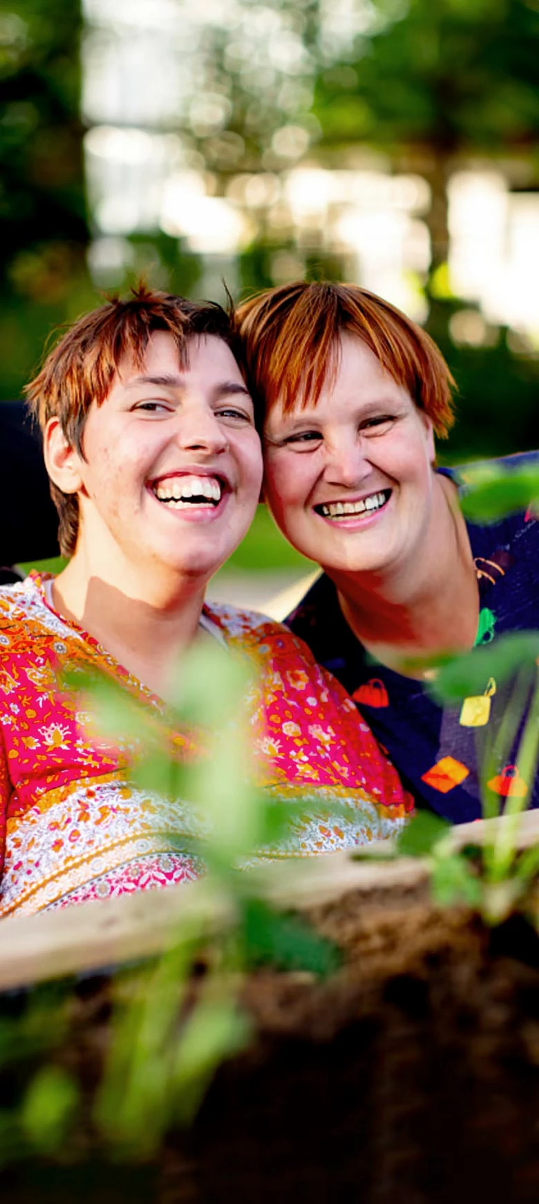 Portrait zweier Frauen in einem grünen Außenbereich. Sie legen ihre Köpfe eng aneinander und lachen in Richtung Kamera. 